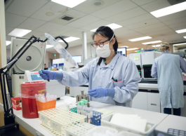 A person wearing glasses, a mask and blue medical protective clothing holds a beaker at a table in a lab.