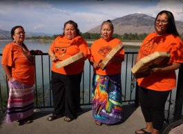 Four Indigenous women wearing orange shirts and two wearing ribbon skirts hold drums and sing on a deck overlooking a lake.