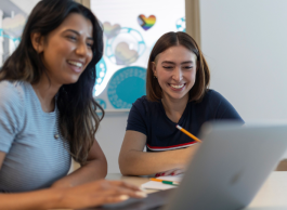 Two smiling young people sit side by side at a table. They're looking at a laptop together.