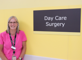A person with medium-length blond-grey hair and glasses wearing a pink t-shirt and lanyard stand in front of a yellow with a sign that say Day Care Surgery.