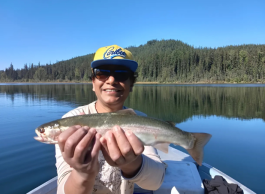 A person with short dark hair wearing sunglasses and a short-sleeved dress shirt poses in front of a lake holding a fish
