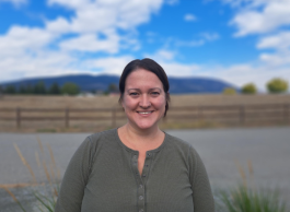 a person wearing a green shirt smiles while standing outdoors by a road and a fence in a farming area