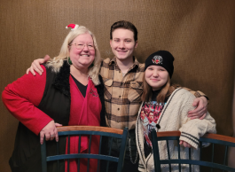 An individual with shoulder length blonde hair, wearing gold rimmed glasses, and a red top is smiling for a photo with their niece and nephew.