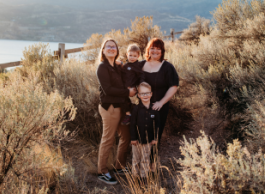 family of four with two small kids smiles to the camera while surrounded by shrubbery outdoors with a lake view
