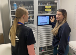 Two people in black t-shirts stand in a medical room. One is showing a monitor to the other.