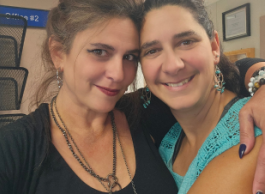 Two people stand arm in arm, heads touching. One wears a black shirt, the other a teal sleeveless sweater.