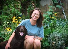 Woman wearing glasses sitting in garden with black dog. Both have big smiles.