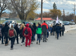 Group of people in warm clothing walking peacefully along a street with a white flag, trees, and parked cars nearby.