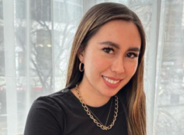 A person with long hair wearing a black top and necklace sitting on a chair with highrises in the background.