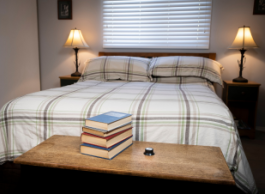 A cozy bedroom with two lamps, a bed with a quilt and books on a chest. A radon detector sits next to the books.