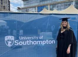 A photo of a woman wearing a black graduation gown and cap standing in front of a fence with a banner that says University of Southampton.