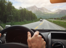 A view of two hands on a steering wheel and dash. Through the window is a highway with forest and mountains in view.