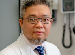 A person with grey hair and beard, wearing glasses, a blue tie and white lab coat stands in front of medical equipment.
