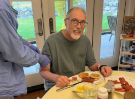 Man with glasses sits at his kitchen table eating breakfast. A health-care worker in blue garments is tying a tourniquet on his arm.