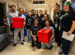 A large group of people stand and kneel in a hospital hallway. Two are carrying big red boxes.