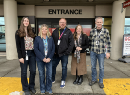 A group of five people in warm clothes and wearing lanyards stand together outside of an emergency department entrance at a hospital.