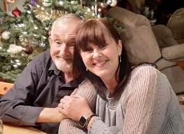Two people smiling near a lit Christmas tree in a cozy, festive room.