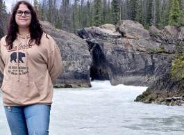 Person standing on rocky riverbank near flowing water with large rock formations and dense evergreen forest in the background.