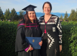 Two people stand in front of a hedge under a blue sky. There are mountains and ocean behind them. One is wearing a graduate cap, and a black and pink robe. Both are wearing Indigenous clothing.