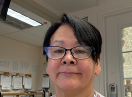 A person with glasses poses in a clinical room with clipboards in the background.