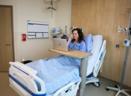 A person in a blue hospital gown wearing headphones sits in a bed watching a laptop on a table.