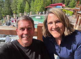 Two smiling people pose for a photo on a deck overlooking a pool on a sunny day.