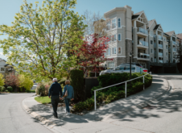 Two older people walk down a sidewalk on a sunny day. Trees and an apartment building can be seen in the background.
