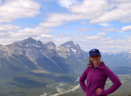 A smiling person wearing a purple coat and blue hat poses on a hill overlooking a valley and mountains.