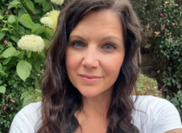 a person with blue eyes and brown hair takes a selfie wearing a white shirt with green shrubbery behind