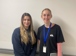 Two young people in casual clothing, one with long blonde hair and one with blonde hair tied back and wearing a lanyard, stand in a beige hallway.