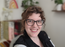 A person wearing black framed glasses and a black vest is shown from the shoulders up facing the camera and smiling in a room with wall art in the background. 