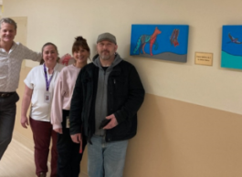 Four people, three in medical attire and one in casual clothes, stand in a hallway next to two bright blue paintings.