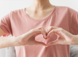 A person wearing a light pink top shown from the neck down holds their hands in the shape of a heart.