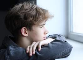 A young person with brown blond hair wearing a grey hoodie looks out a window, their arms crossed in front of them.