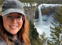 A person wearing glasses and hat poses for a picture by a cliff with a waterfall.