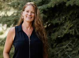A person with long blonde hair wearing a black sleeveless top with a zipper stands in front of a pine tree.