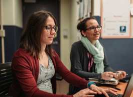 Two people in casual clothing sit together at a desk looking at a computer.