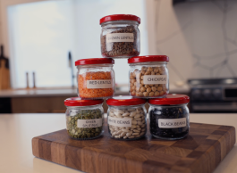 A stack of six glass jars labelled with various kinds of beans sit on a wooden breadboard on a kitchen counter.