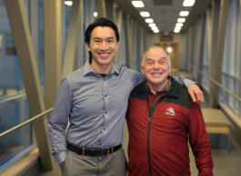 Two smiling people stand arm in arm inside a bright catwalk. One is wearing a dress shirt, the other a red and black jacket.