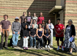 A large group of youth, some sitting, some standing, pose in front of a brick wall on a sunny day.