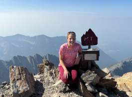A person in a pink shirt kneeling down next to a sign on a mountain top.