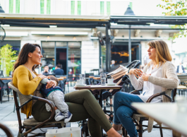 Two people sit at a table at an outdoor cafe. An older person is speaking to a younger person who is breastfeeding their baby.