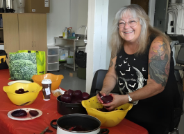 A person with long grey hair and glasses, with an arm tattoo, peels beets at a table covered in a red tablecloth.