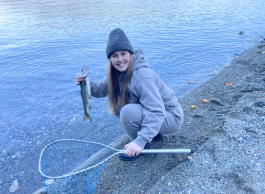 Person crouching on a rocky shoreline holding a freshly caught fish, with a fishing net beside the water.