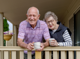 An older couple lean against a railing on a wooden porch. Both are holding coffee cups.