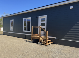 A long dark grey building with white framed windows and doors. A small wooden step leads up to the door.