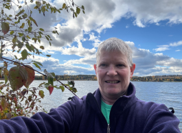 A person with short grey hair takes a selfie in front of a lake surrounded by low mountains on a partly cloudy day.