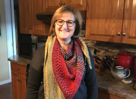 A person with short brown hair and bangs wearing glasses with a red and gold knitted scarf stands in a kitchen with brown cupboards.