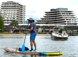 A person paddleboards on calm water near a boat, with modern waterfront buildings in the background.