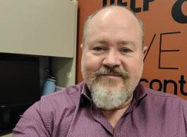 Person with a short beard wearing a purple button-up shirt, seated at a desk in an office with a computer monitor and orange wall signage in the background.